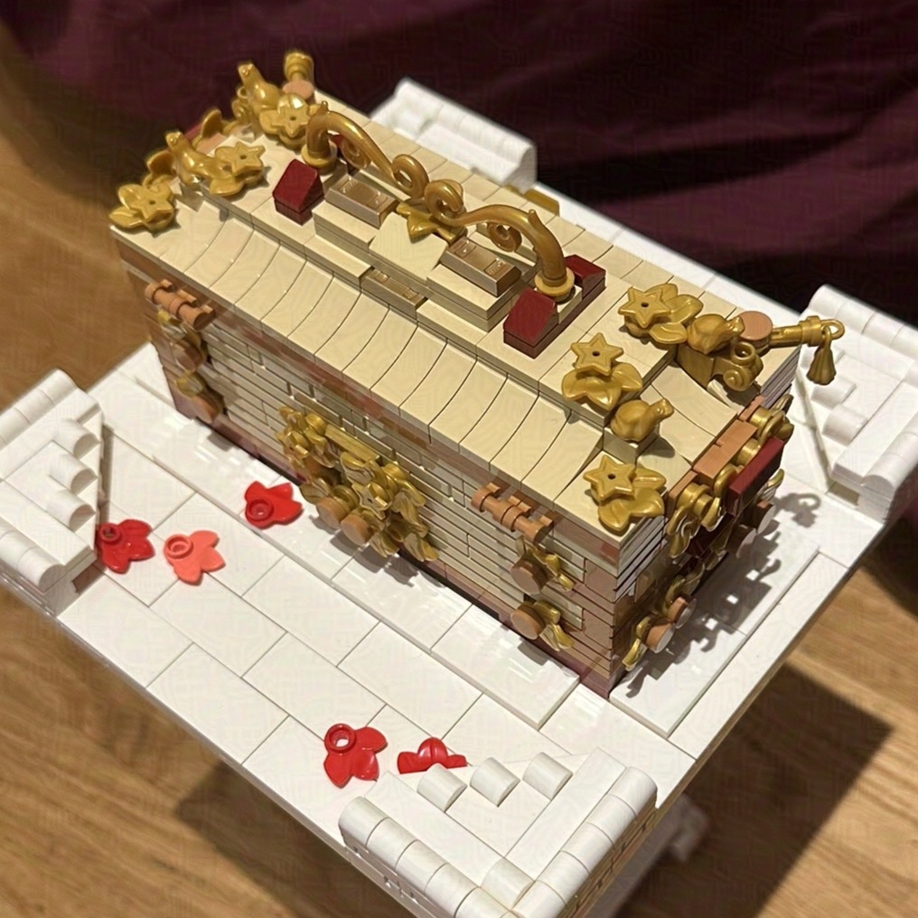 A LEGO model of a decorative hope chest with ornate gold and dark red accents, set atop a white, block-built base. The chest is rectangular with tan brick detailing, a gold filigree design, and dark red accent pieces. Small red rose-like petal LEGO pieces are scattered on the base. The scene is lit, with a wooden floor and burgundy cloth backdrop.